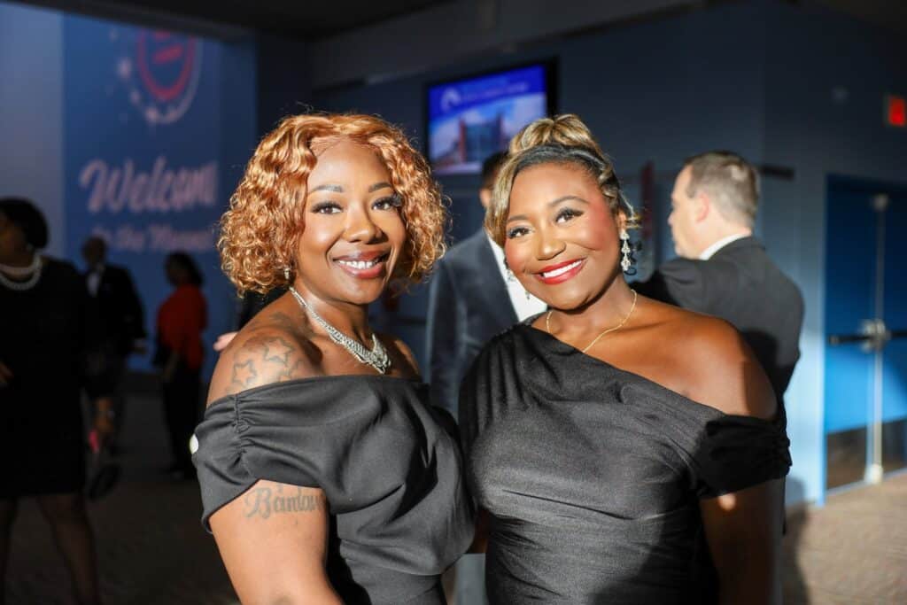 654854997_1358081286353196_276897606401225057_n-1024x683 | Akron Urban League Two Women in Off-shoulder Black Dresses Pose and Smile at a Formal Event, with a Blue Backdrop in the Background.