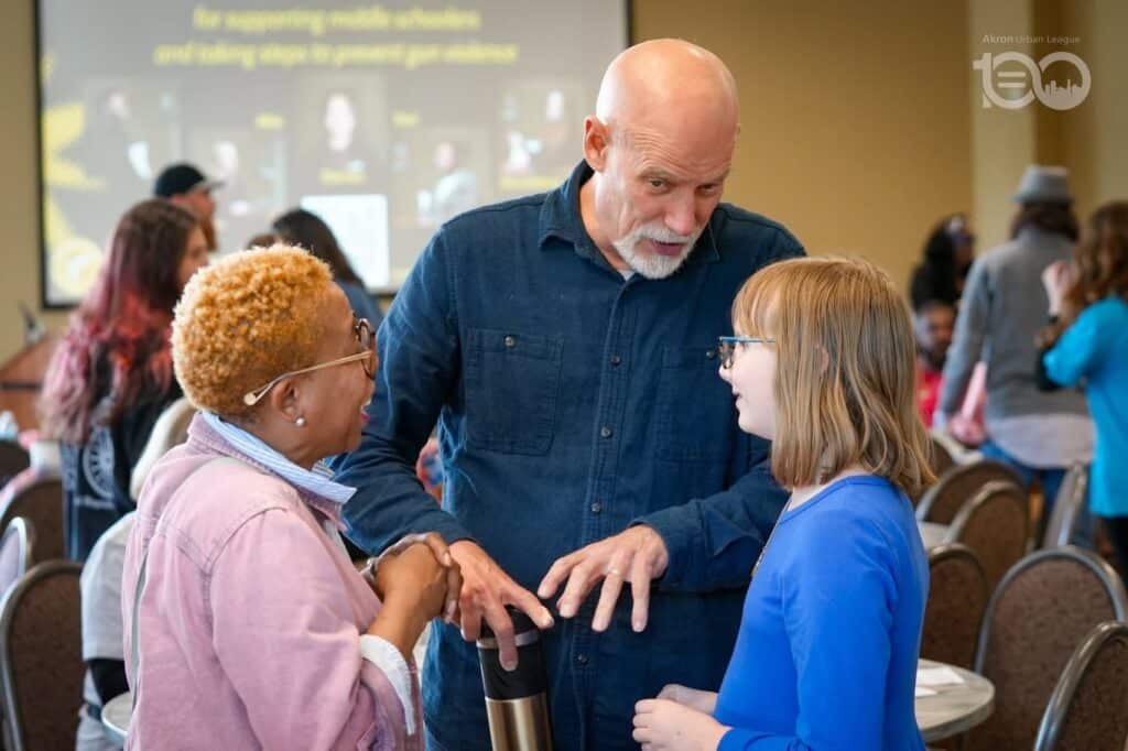 686514438_18226207360314357_8518069740026835894_n-1024x682 | Akron Urban League Older Man with a Beard in a Blue Shirt Talks with a Young Girl and a Woman, Their Hands Meeting in a Friendly Handshake at a Crowded Event Venue.