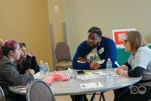 686902465_18226207339314357_6416585425757379443_n-600x400 | Akron Urban League Five People Sit Around a Round Table in a Discussion, with Papers, Name Tags and Water Bottles on the Table in a Bright Room.