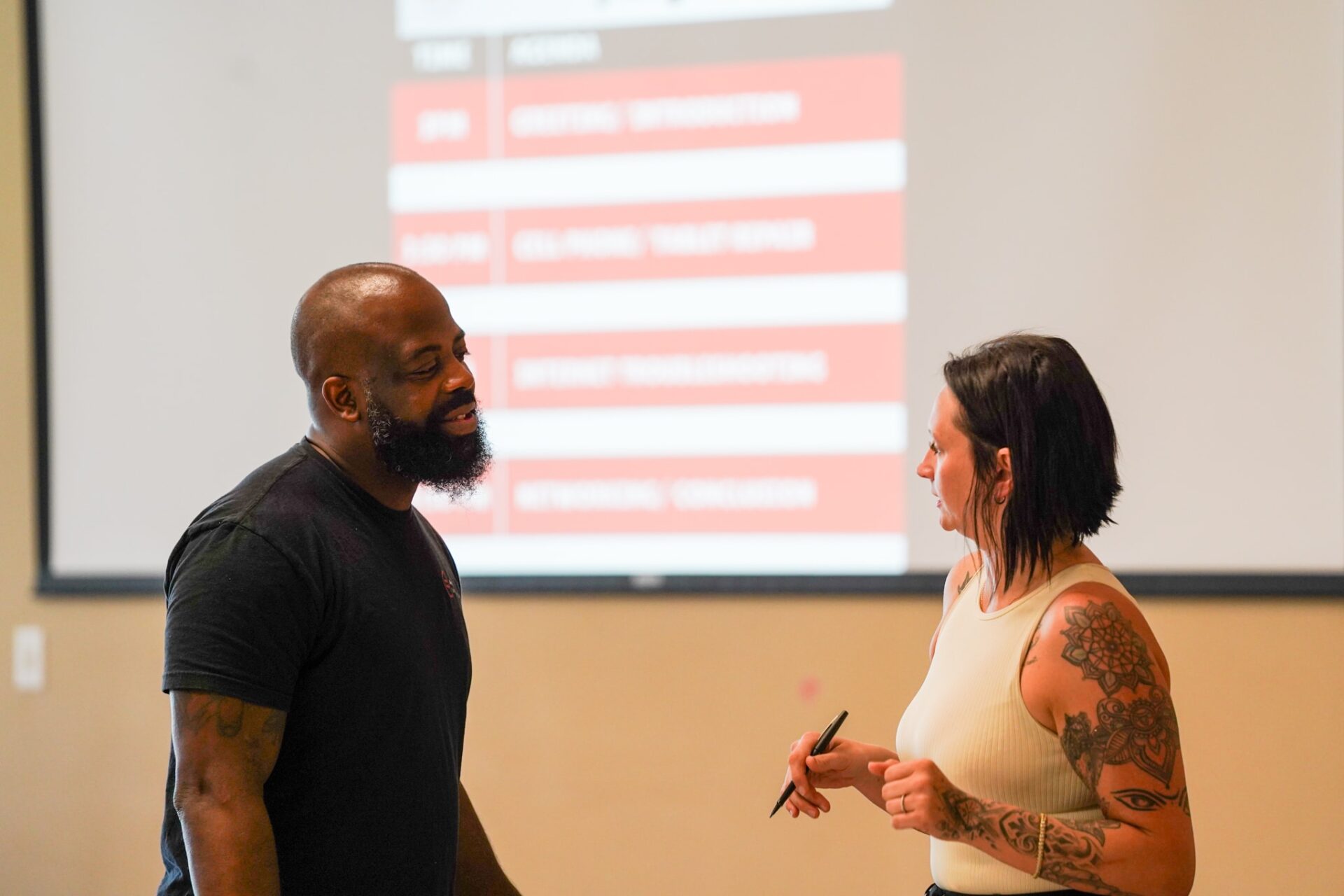 Anti-recidivism | Akron Urban League Bald Man with Beard in a Black Shirt Speaks to a Woman with Arm Tattoos Holding a Pen, Near a Projected Slide.