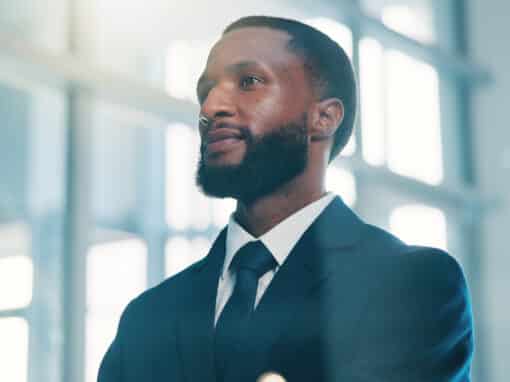 Confident-businessman-in-a-suit-looking-thoughtful-2026-06-08-17-44-38-utc Portrait of a bearded man in a dark suit and tie, looking upward in a bright, modern office hallway