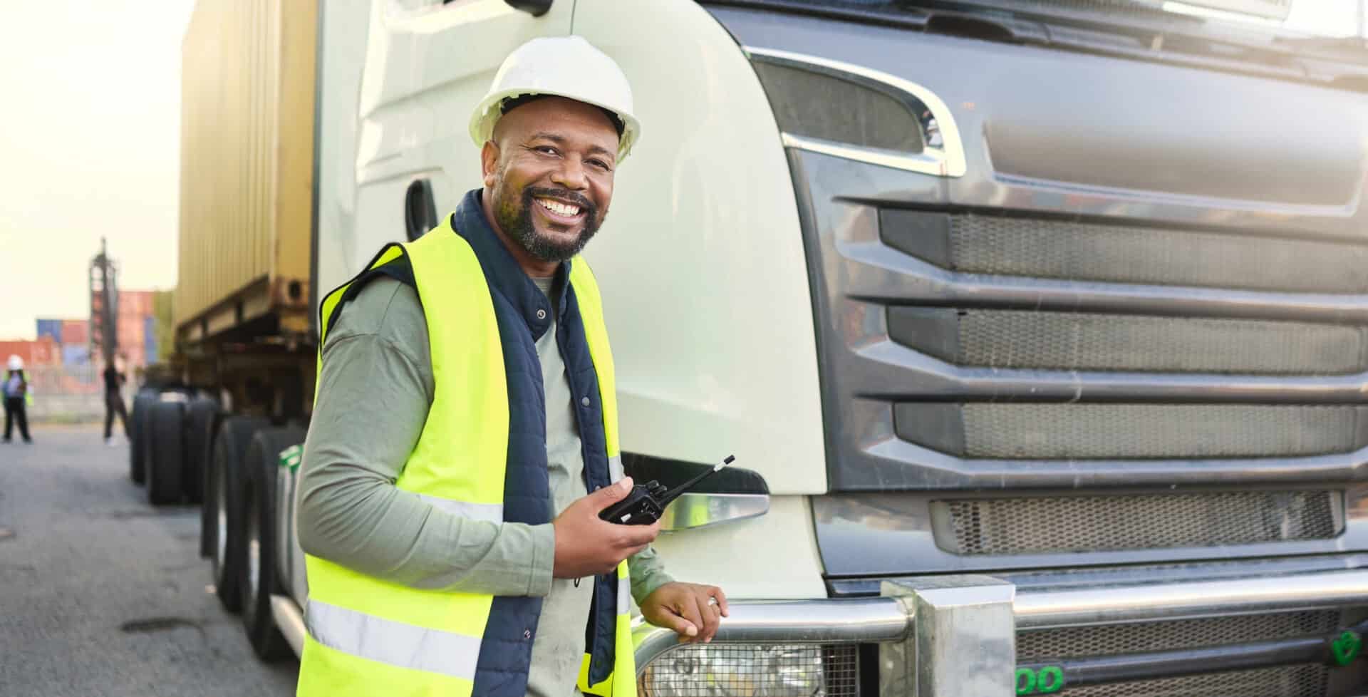 Construction-worker-in-shipping-business-by-delive-2023-11-27-04-56-13-utc | Akron Urban League Smiling Truck Driver in a Neon Safety Vest and Hard Hat Standing Beside a Large White Semi-truck, Holding a Radio.