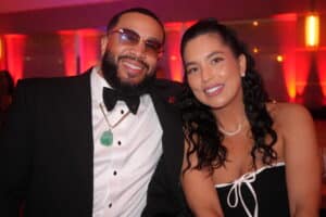 Dsc05335-300x200 | Akron Urban League Smiling Couple Posing for a Photo at a Formal Event; Man in Tuxedo with Bow Tie and Sunglasses, Woman in a Black Strapless Dress with White Trim and Long Earrings, Red Ambient Lighting Behind Them.