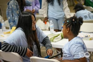Two Women in Light Blue Shirts Chat and Smile at a Table During a Community Event, with a Young Girl with Braided Hair Nearby.