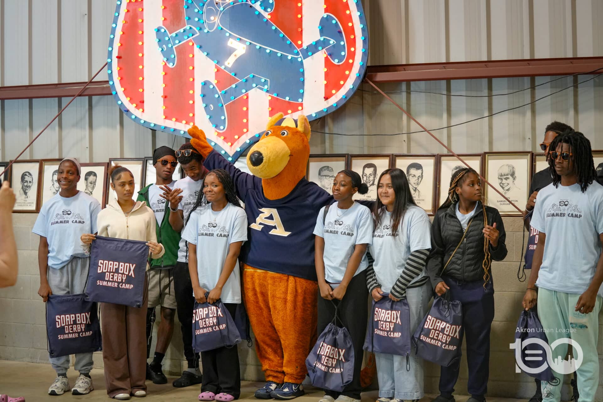 Group of Campers in Light Blue Shirts Posing with a Large Orange Bear Mascot Under a Neon Sign, Each Holding Soapbox Derby Summer Camp Bags in a Gym Hall Setting.