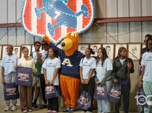 Group of campers in light blue shirts posing with a large orange bear mascot under a neon sign, each holding Soapbox Derby Summer Camp bags in a gym hall setting.