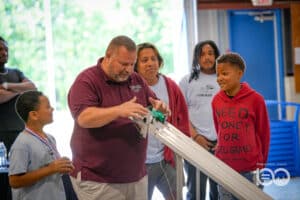 Dsc06335 | Akron Urban League Man in Maroon Polo Explains a Small Model Launch Ramp to Two Boys While Others Watch at a Community Science Event.