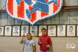 Two Smiling Young Athletes Pose with Trophies After a Competition, Medals Around Their Necks, in Front of Wall of Framed Portraits Under a Bright Neon Sign. Logo Reads Akron Urban League 100 at Corner.
