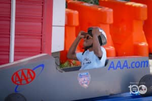Person Wearing a Helmet and Orange-tinted Sunglasses Sits in a Gray Soapbox Derby Car, with Bright Orange Barriers Behind Them and Aaa Branding on the Car.