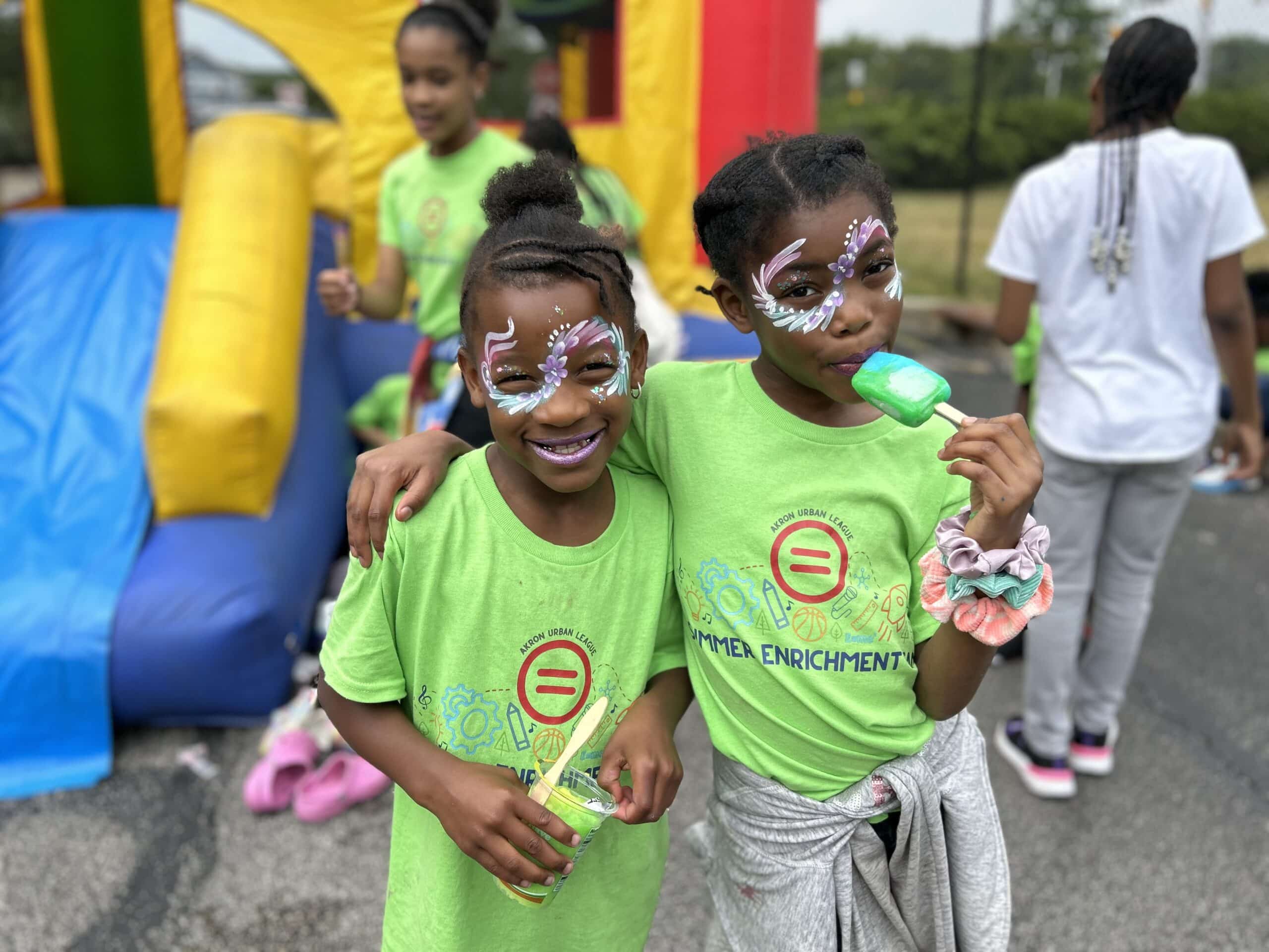 Education & Youth | Akron Urban League Two Children with Bright Face Paint and Green Shirts Pose with Popsicles in Front of a Yellow and Red Bouncy House.