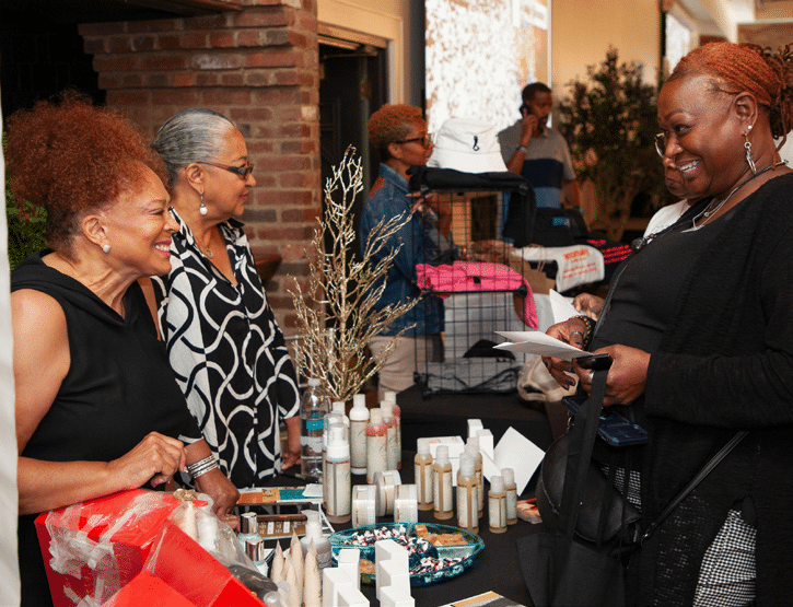 Entrepreneurship | Akron Urban League Three Women Smile over a Table Display of Beauty Products in Bottles and Jars at an Indoor Pop-up Market.