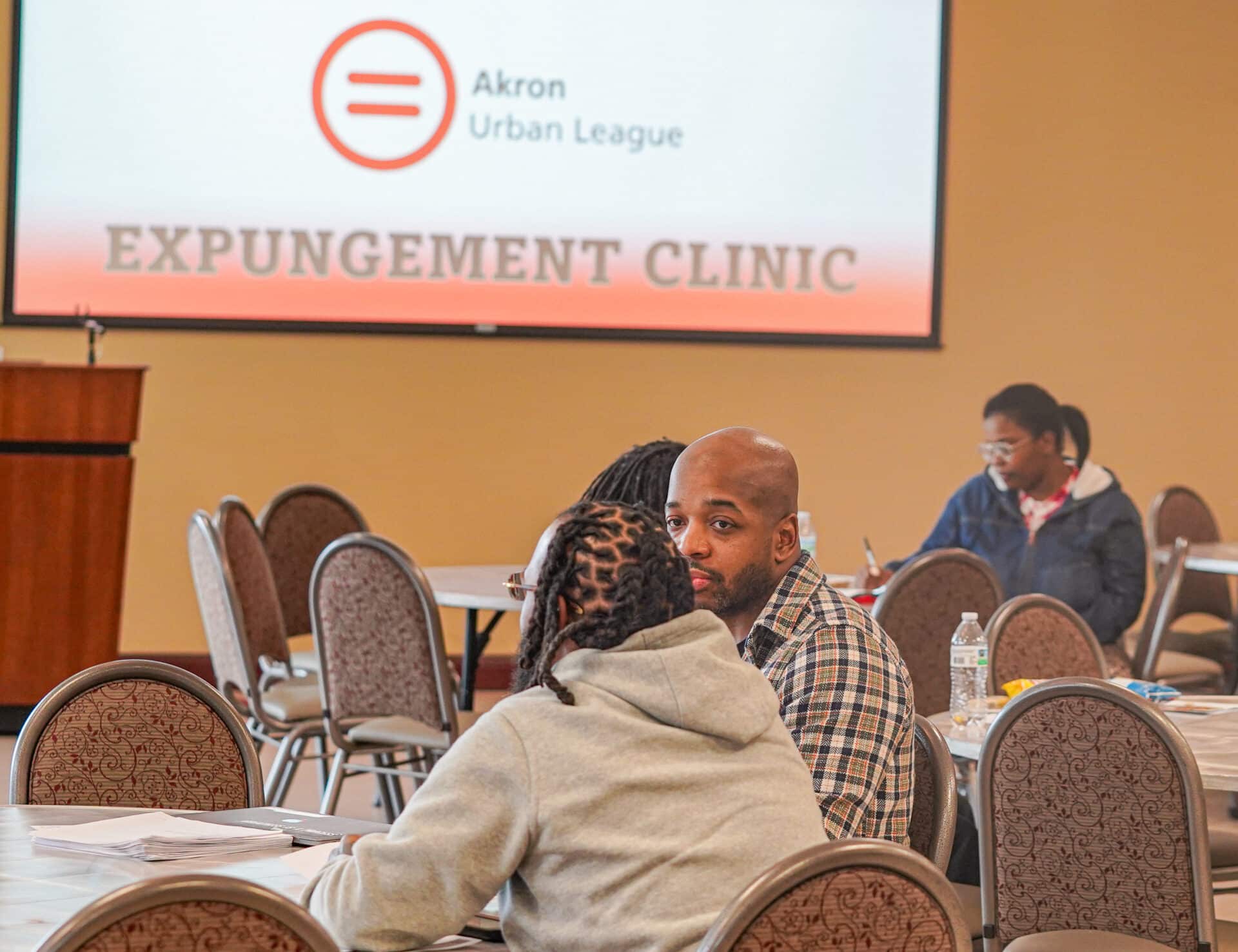 Expungement | Akron Urban League Group of People Seated at Round Tables in a Clinic Event, with a Banner Reading 'expungement Clinic' in the Background and Akron Urban League Logo Above It.