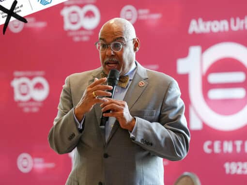 Flyers Blog Black man in a gray suit and bow tie speaks into a microphone on stage at a Centennial celebration backdrop that reads Akron Urban League.
