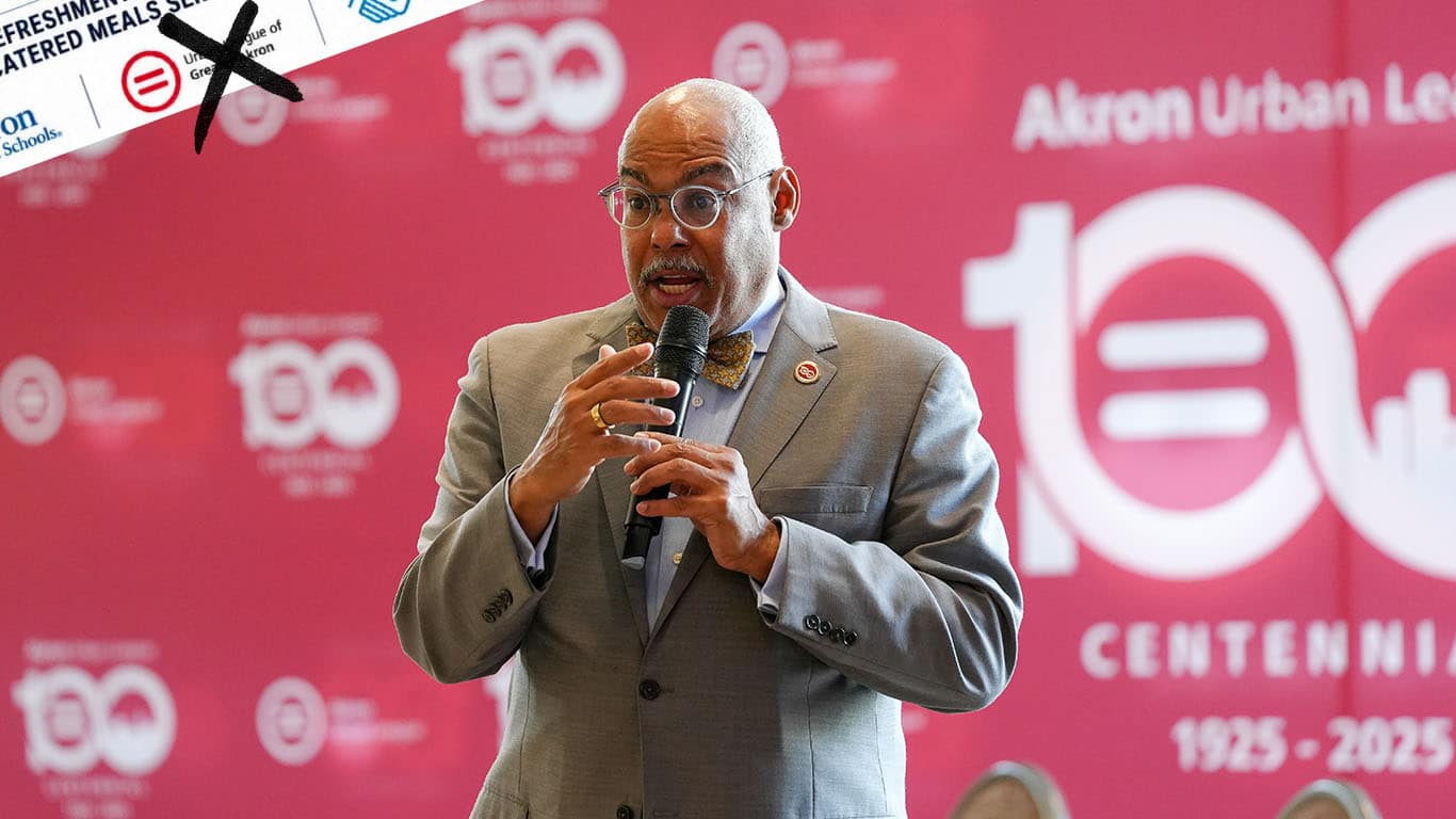 Black Man in a Gray Suit and Bow Tie Speaks into a Microphone on Stage at a Centennial Celebration Backdrop That Reads Akron Urban League.