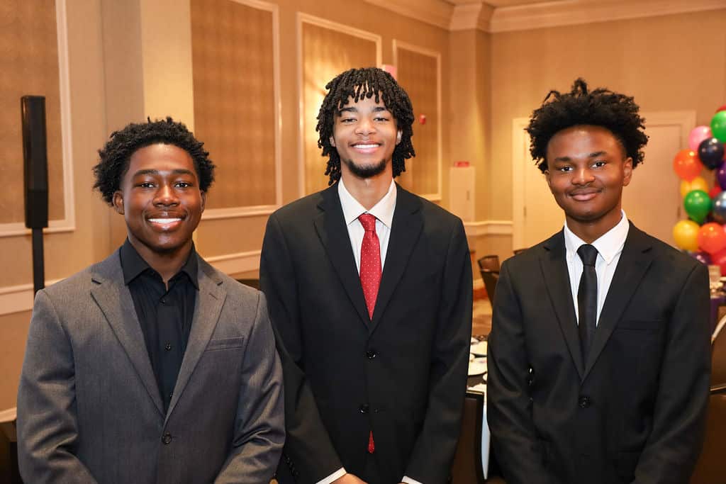 Img_0706 | Akron Urban League Three Young Men in Suits Posing and Smiling at a Formal Event in a Banquet Hall with Colorful Balloons in the Background.