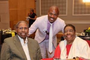 Img_0723-300x200 | Akron Urban League Three Smiling Adults at a Banquet Table in a Conference Room; a Man in a Light Pink Blazer Stands Behind Two Seated Attendees.