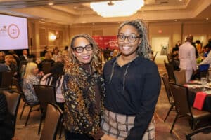 Img_0766-300x200 | Akron Urban League Two Women Smile for the Camera at a Conference or Networking Event, with Attendees and Round Tables in the Background.