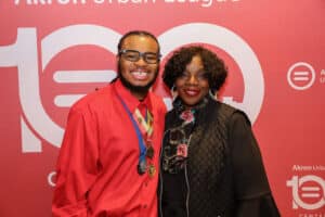Img_0768-300x200 | Akron Urban League Two People Smiling for a Photo at a Red Event Backdrop with Branding; the Man Wears a Red Shirt and a Medal on a Blue Ribbon, the Woman Wears Black.