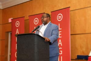 Img_0772-300x200 | Akron Urban League Male Speaker in a Blue Suit Delivering a Keynote at a Podium, with Red Event Banners in the Background.