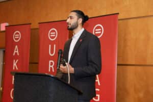 Man in a Dark Suit Giving a Speech at a Podium with Red Banners Behind Him.]','