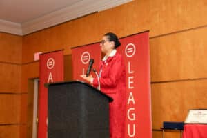 Img_0796-300x200 | Akron Urban League Woman in a Red Blazer Speaks at a Black Podium, Microphone in Hand, with Red Banners Behind in a Wood-paneled Room.