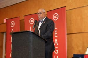 Img_0806-300x200 | Akron Urban League Older Man in a Dark Suit Delivering a Speech at a Lectern, Against Red Banners with White Circular Logos Behind Him.