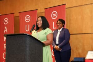 Img_0818-300x200 | Akron Urban League Woman in a Light-green Dress Speaks at a Lectern on Stage, with Another Woman in a Blue Blazer Standing Nearby As Red Banners Hang Behind.