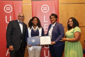 Img_0826-300x200 | Akron Urban League Four Adults on Stage at an Award Ceremony; a Recipient Holds a Certificate As a Man Presents It, with Two Others Smiling in Front of Red Banners.