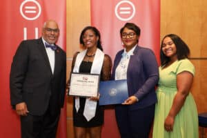 Img_0835-300x200 | Akron Urban League Four Professionals Posing on Stage at an Awards Event; One Woman Holds a Certificate and Another Holds a Blue Folder, with Red Banners in the Background.