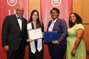 Img_0856-300x200 | Akron Urban League Group Portrait of Four Adults at an Awards Ceremony: a Man in a Suit, a Student Holding a Certificate, a Woman with a Blue Folder, and Another Woman in a Green Dress, with Red Banners Behind.