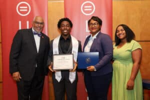 Img_0863-300x200 | Akron Urban League Four Adults Posing at an Award Ceremony; the Recipient in the Center Holds a Certificate, Others Smile Beside Him with Red Banners Behind Them.
