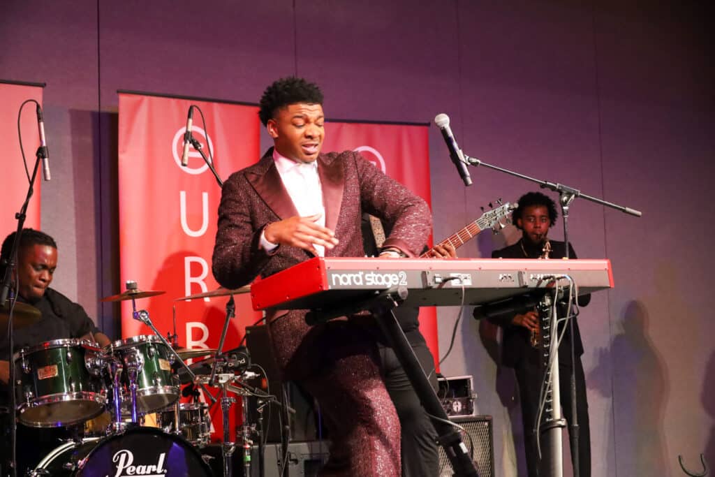Img_2473-1024x683 | Akron Urban League Male Musician in a Maroon Glitter Suit Plays a Red Nord Keyboard on Stage, with a Drummer and Saxophonist in the Background and Red Banners Behind.