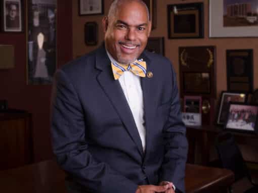 John Headshot Smiling man in a navy suit and yellow striped bow tie sits in an office lined with awards and photos on the wall in the background.