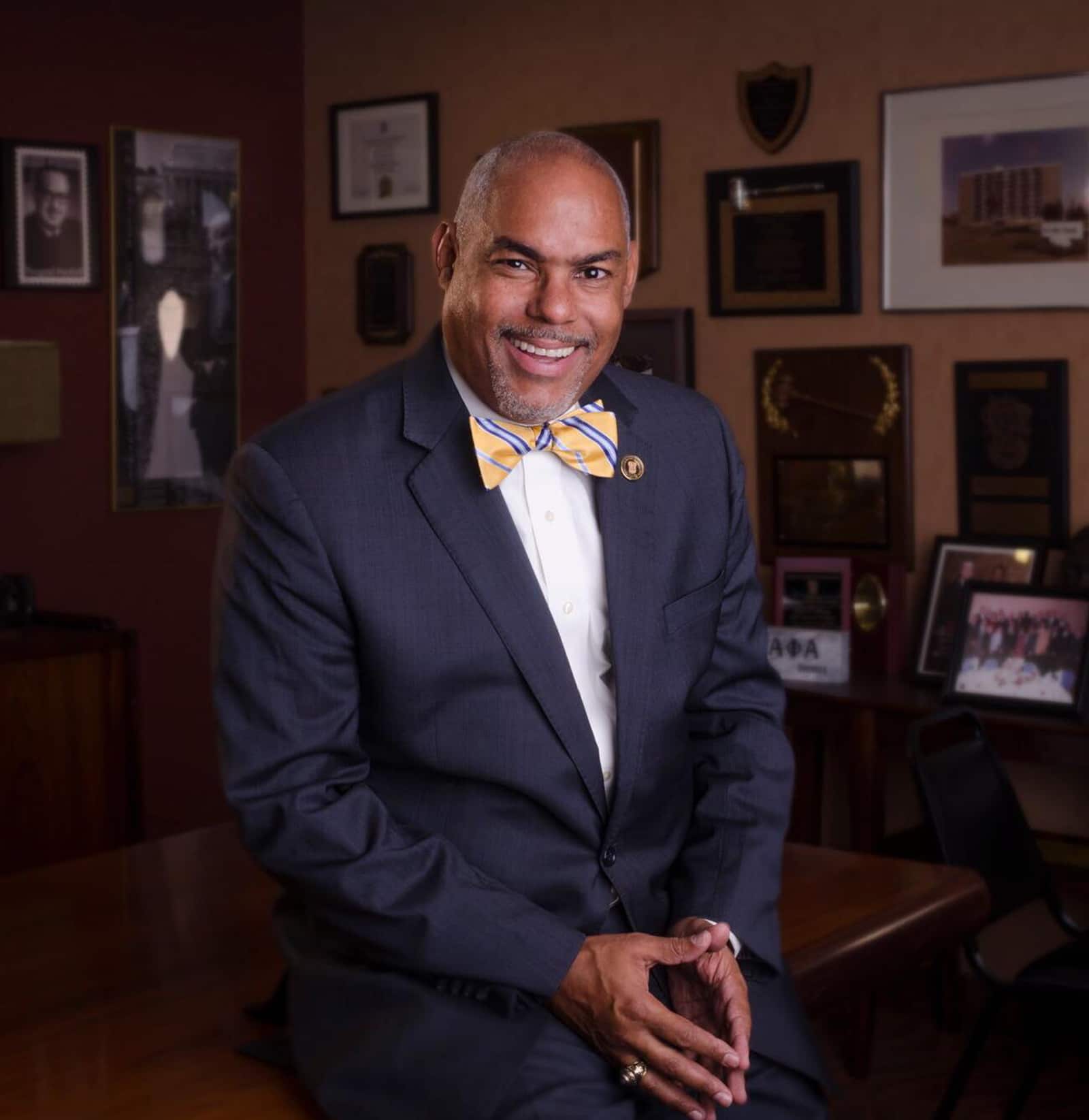 John Headshot | Akron Urban League Smiling Man in a Navy Suit and Yellow Striped Bow Tie Sits in an Office Lined with Awards and Photos on the Wall in the Background.