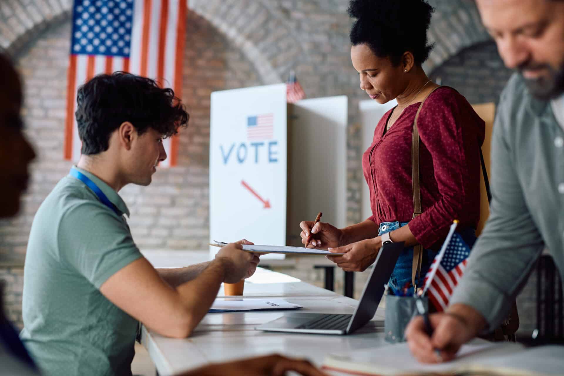 Election Volunteers Assist a Voter at a Registration Table with a Clipboard, Us Flag in the Background.