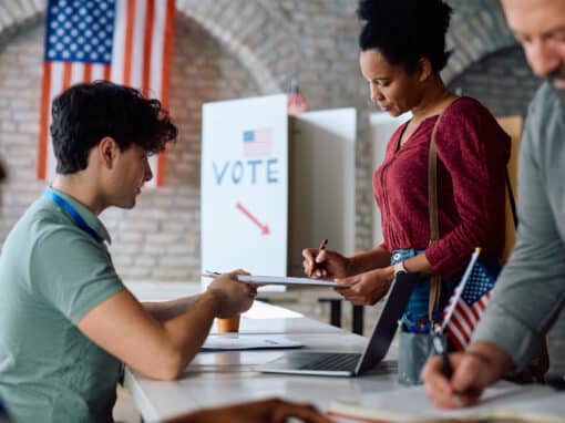 Black Woman Signing Up to Vote at Polling Place During Us Elections Election volunteers assist a voter at a registration table with a clipboard, US flag in the background.