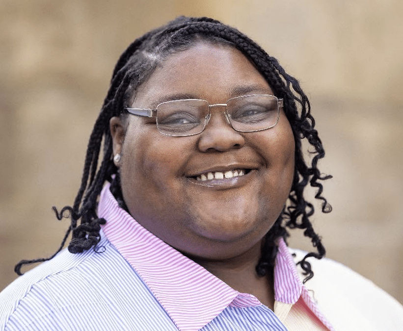 Smiling woman with glasses and braided hair wearing a pink-striped collared shirt.