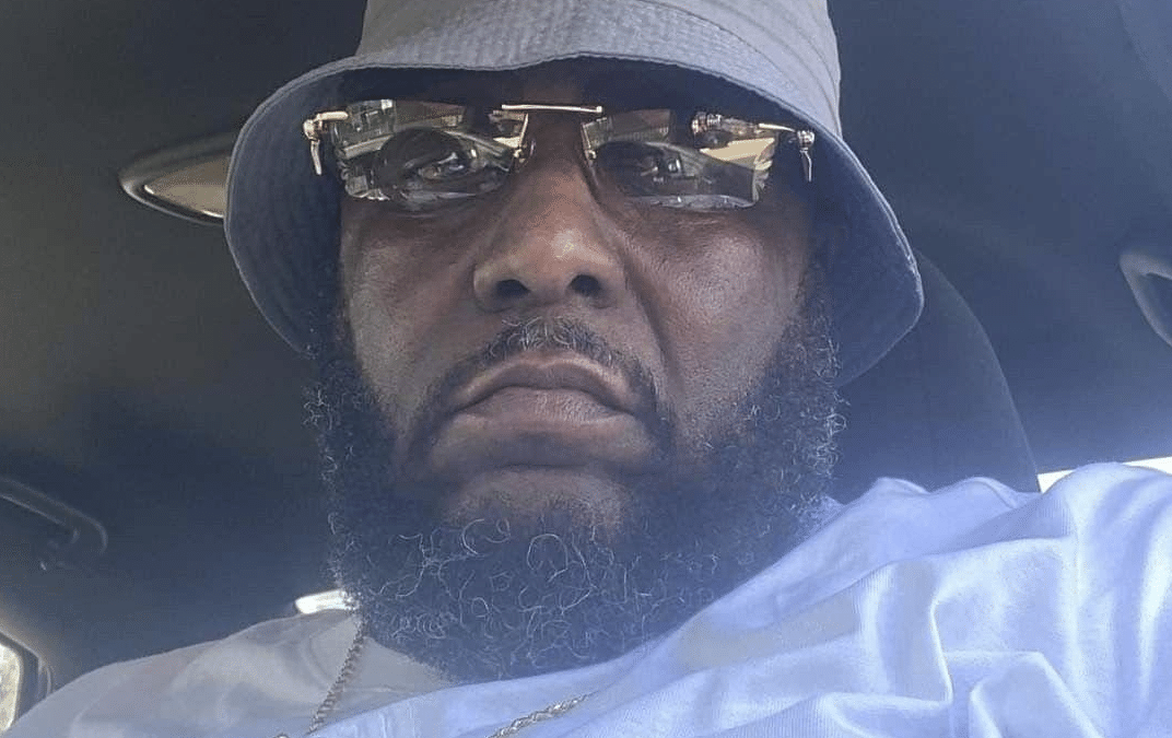 Man in a car wearing a gray bucket hat, mirrored sunglasses, and a gold chain with a light shirt.