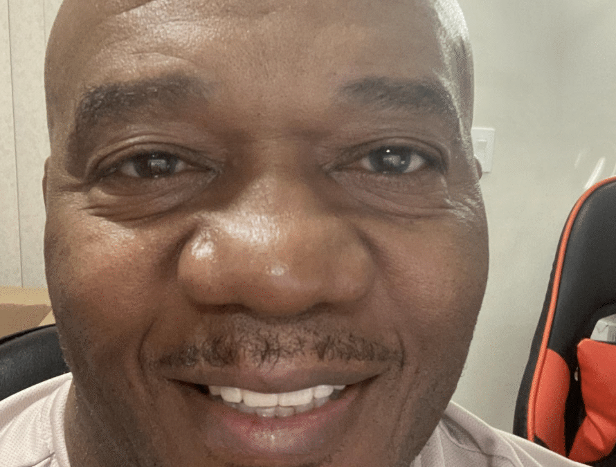 Close-up of a smiling Black man with a bald head, wearing a light shirt indoors.