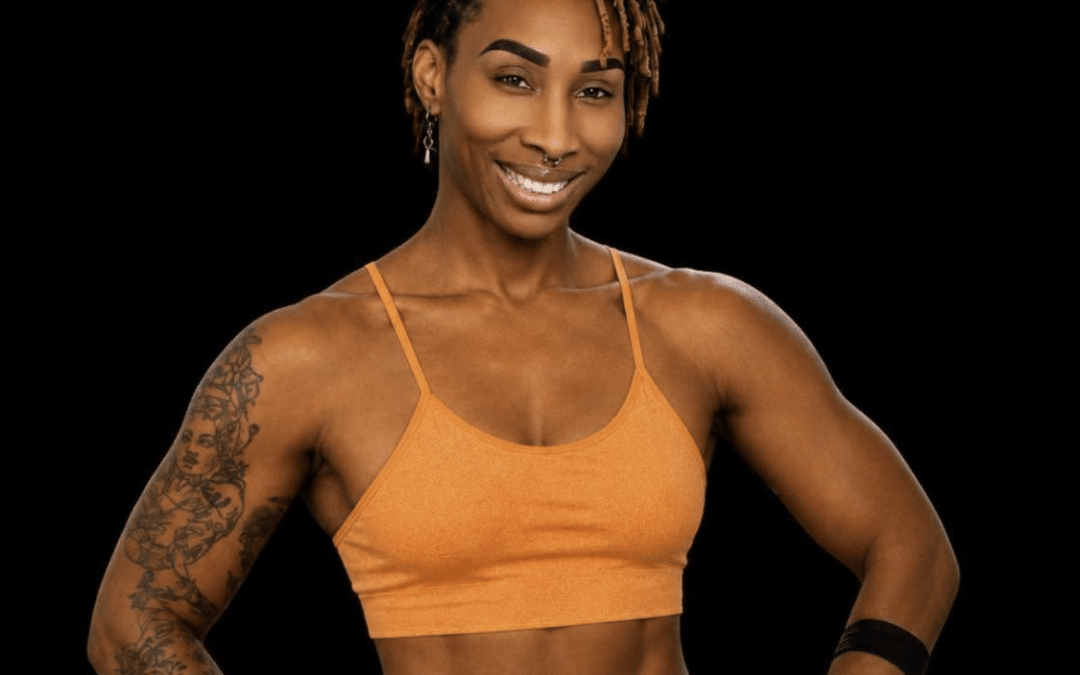 Smiling Black woman in an orange sports bra and matching bottoms, posing with hands on hips against a black background to highlight her athletic build.