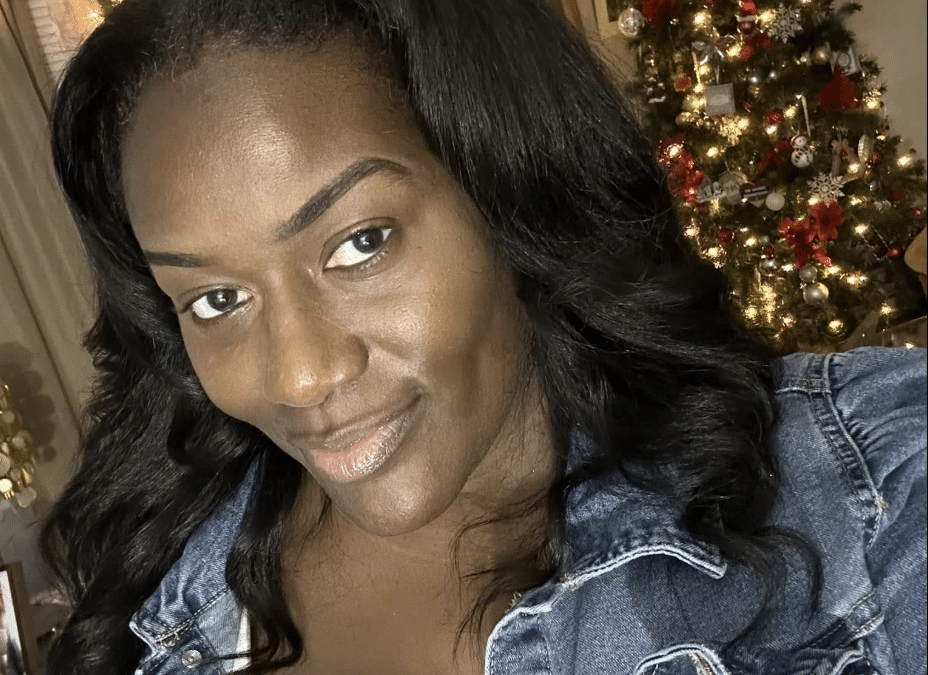 Woman taking a selfie indoors, wearing a denim jacket, with a lit Christmas tree in the background.