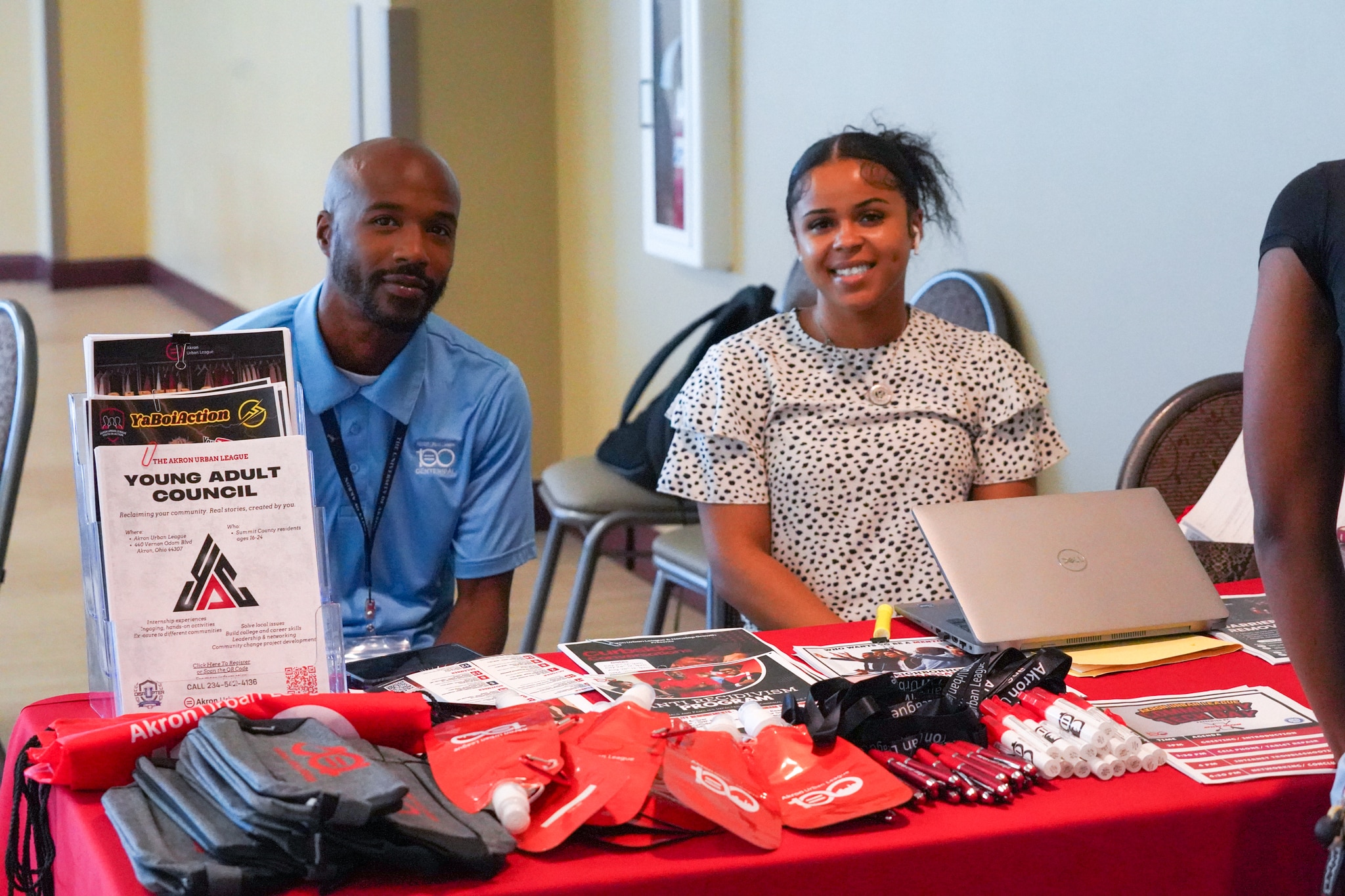 Workforce Success Center | Akron Urban League Two Young Adults Smiling Behind a Table with Flyers, Pens, and Red Swag, Including One Titled "young Adult Council."